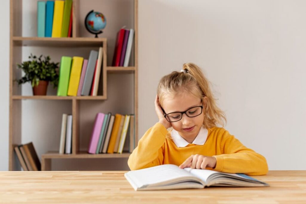 a girl with glasses and orange sweater reading book sitting on the table